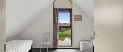 A bedroom with a sloped ceiling, a bed, a chair, a nightstand, and a window with a view of a house and greenery.