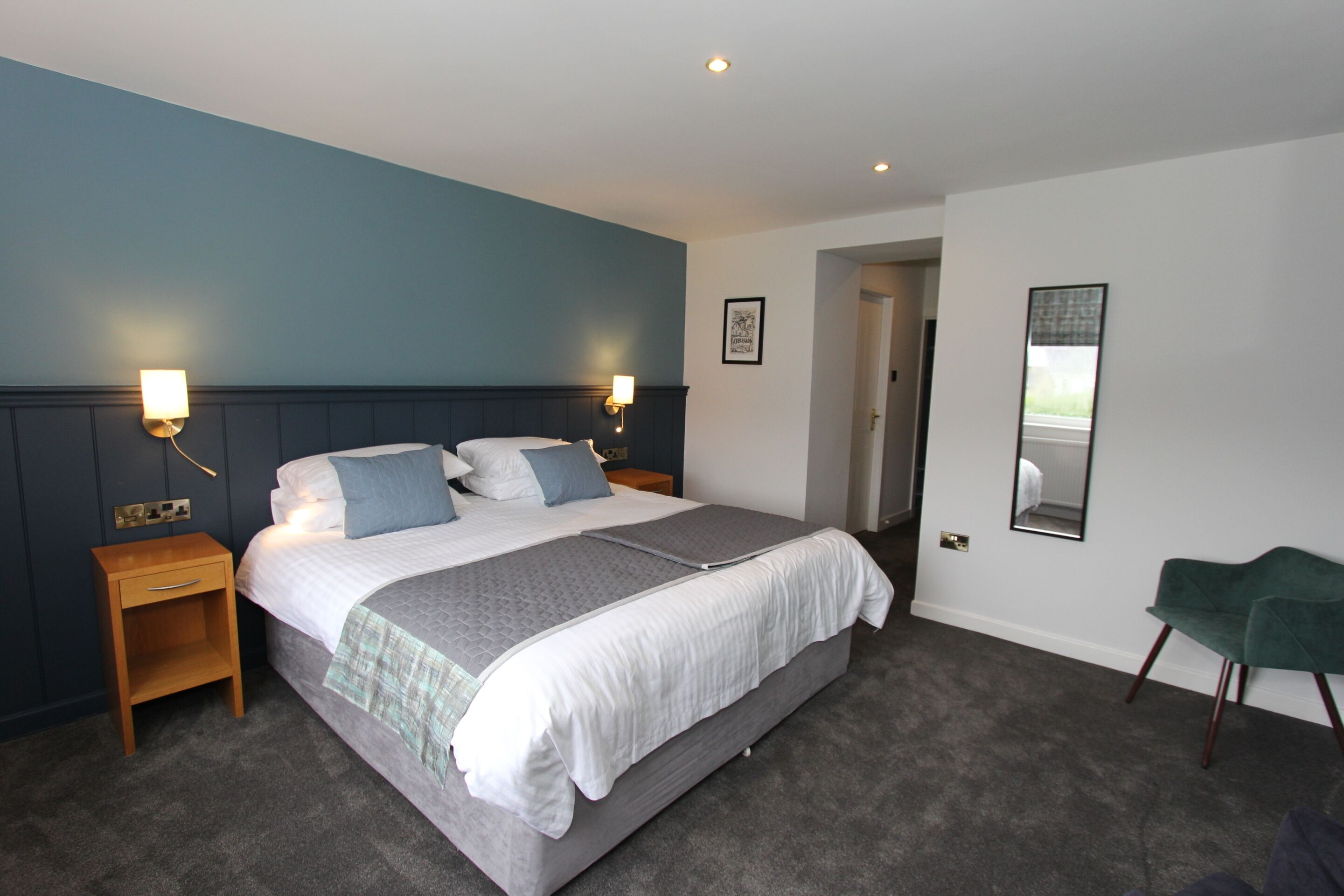 A modern bedroom with a large bed, bedside tables, a green chair, and a door leading to another room.