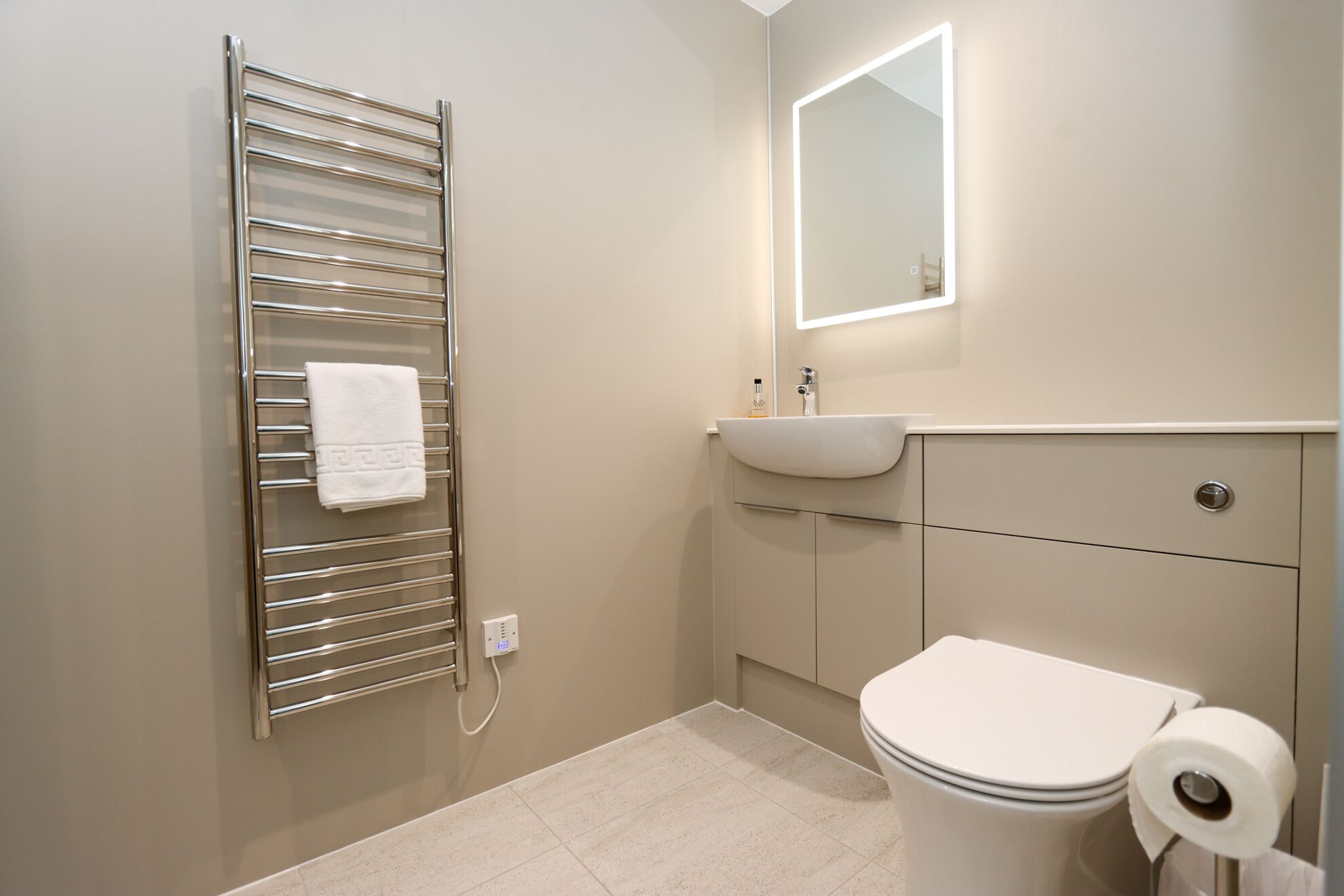 Een moderne badkamer met een chromen handdoekrek, een witte wastafel, een toilet en een spiegel met verlichting.