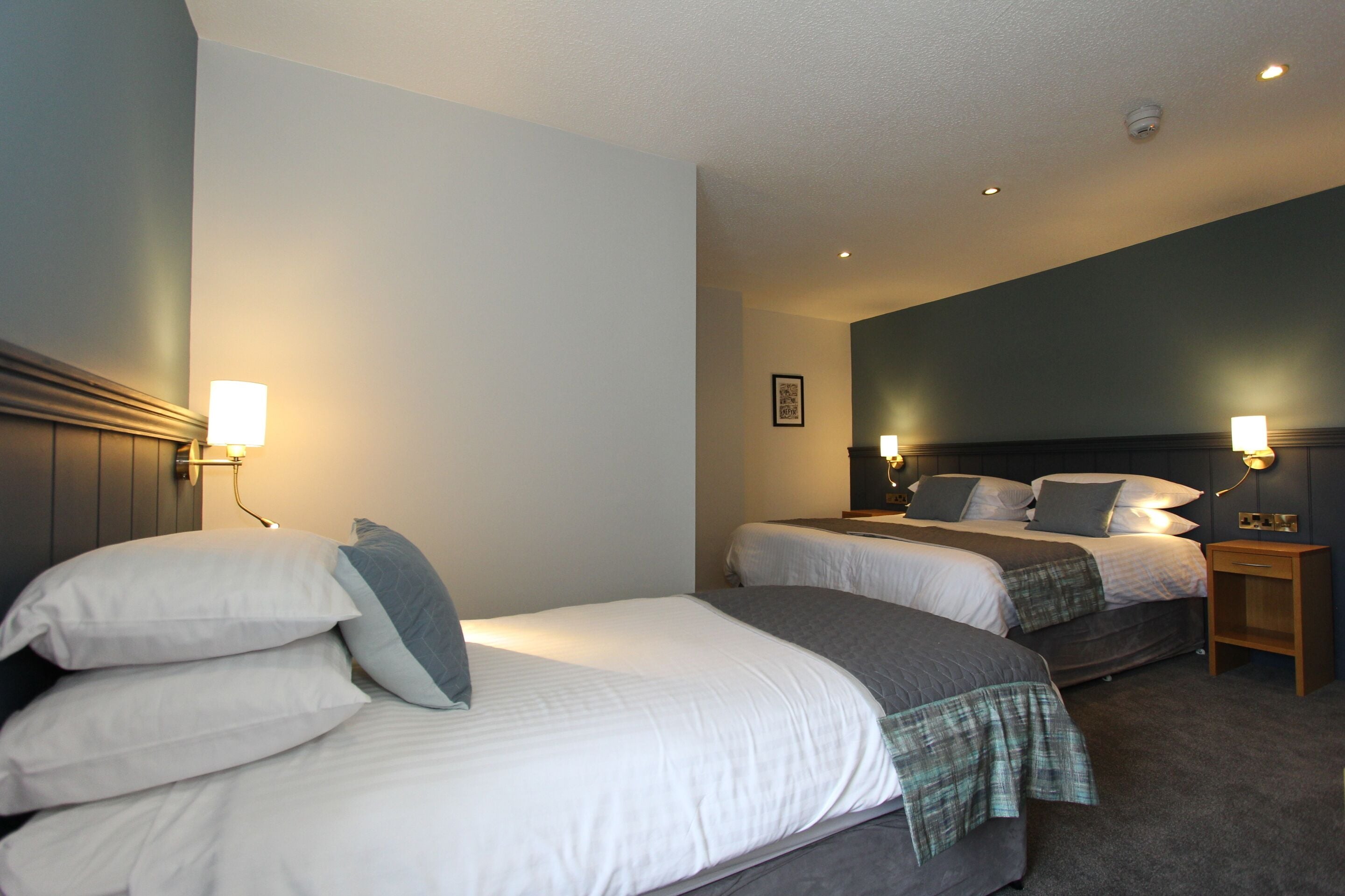 A hotel room with two beds, each with white linens and gray bedding, and a small wooden nightstand with a lamp.