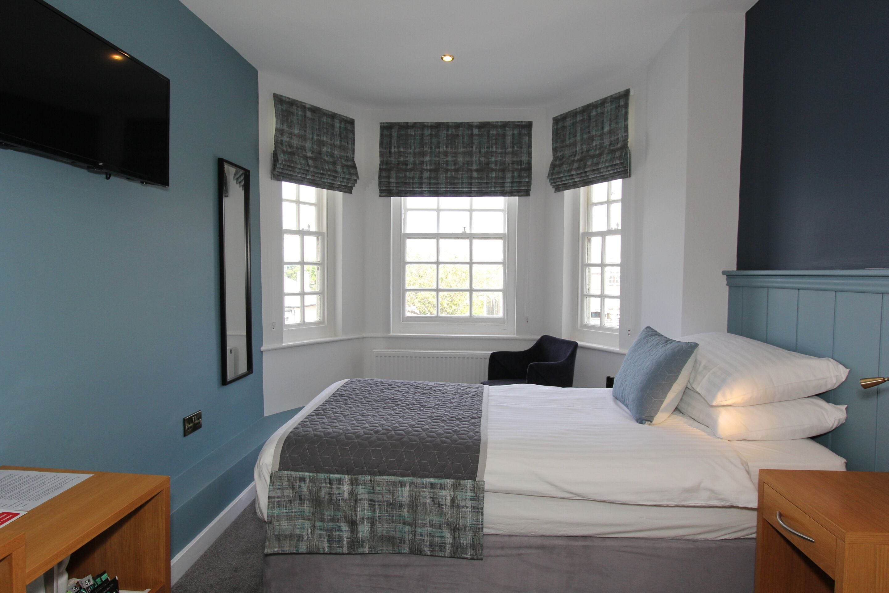 A bedroom with a bed, a desk, and a chair. There are windows with curtains and a television on the wall.