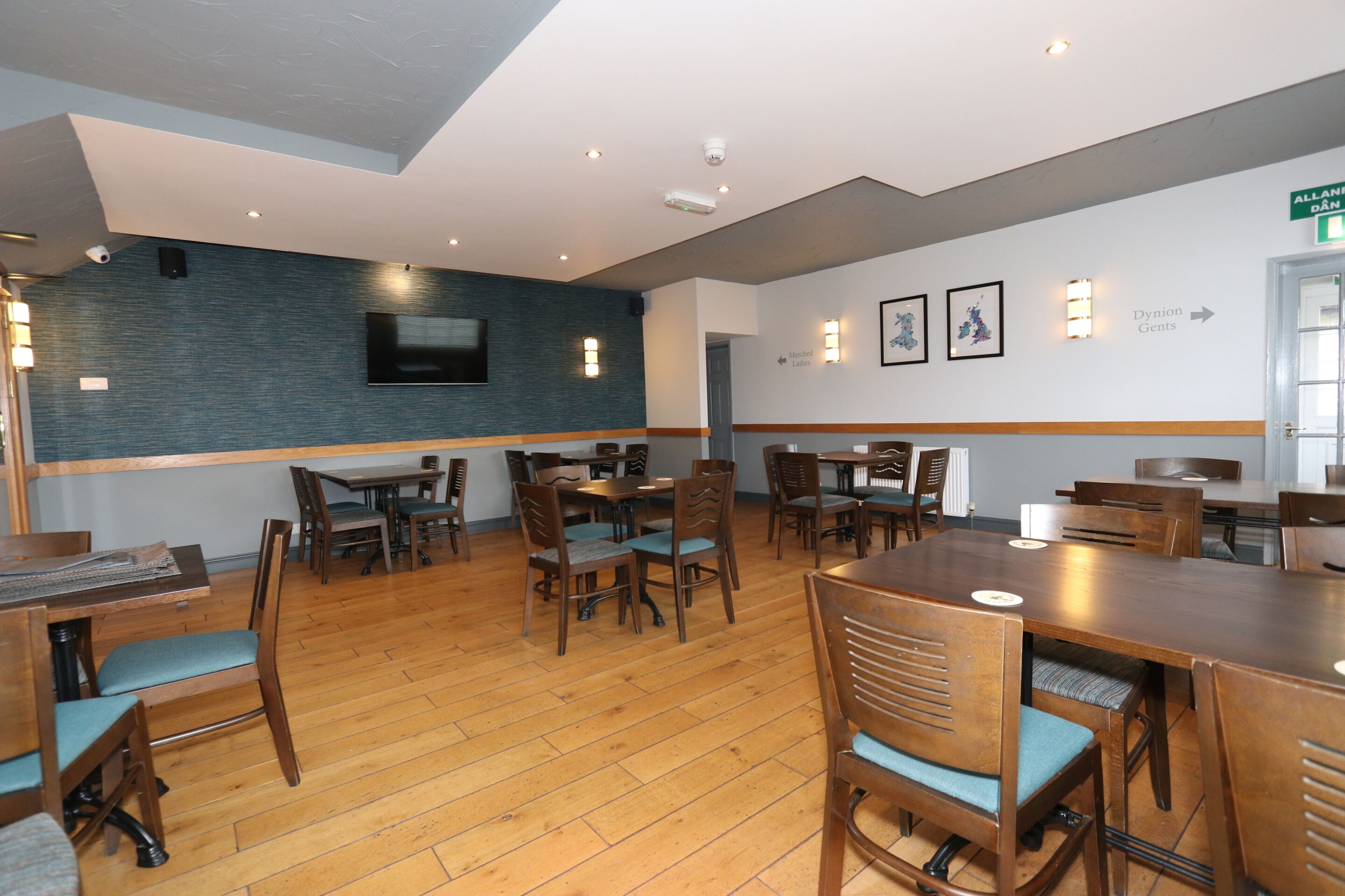 A hotel dining area with wooden tables and chairs, a brick accent wall, and a flat-screen TV mounted above the seating.