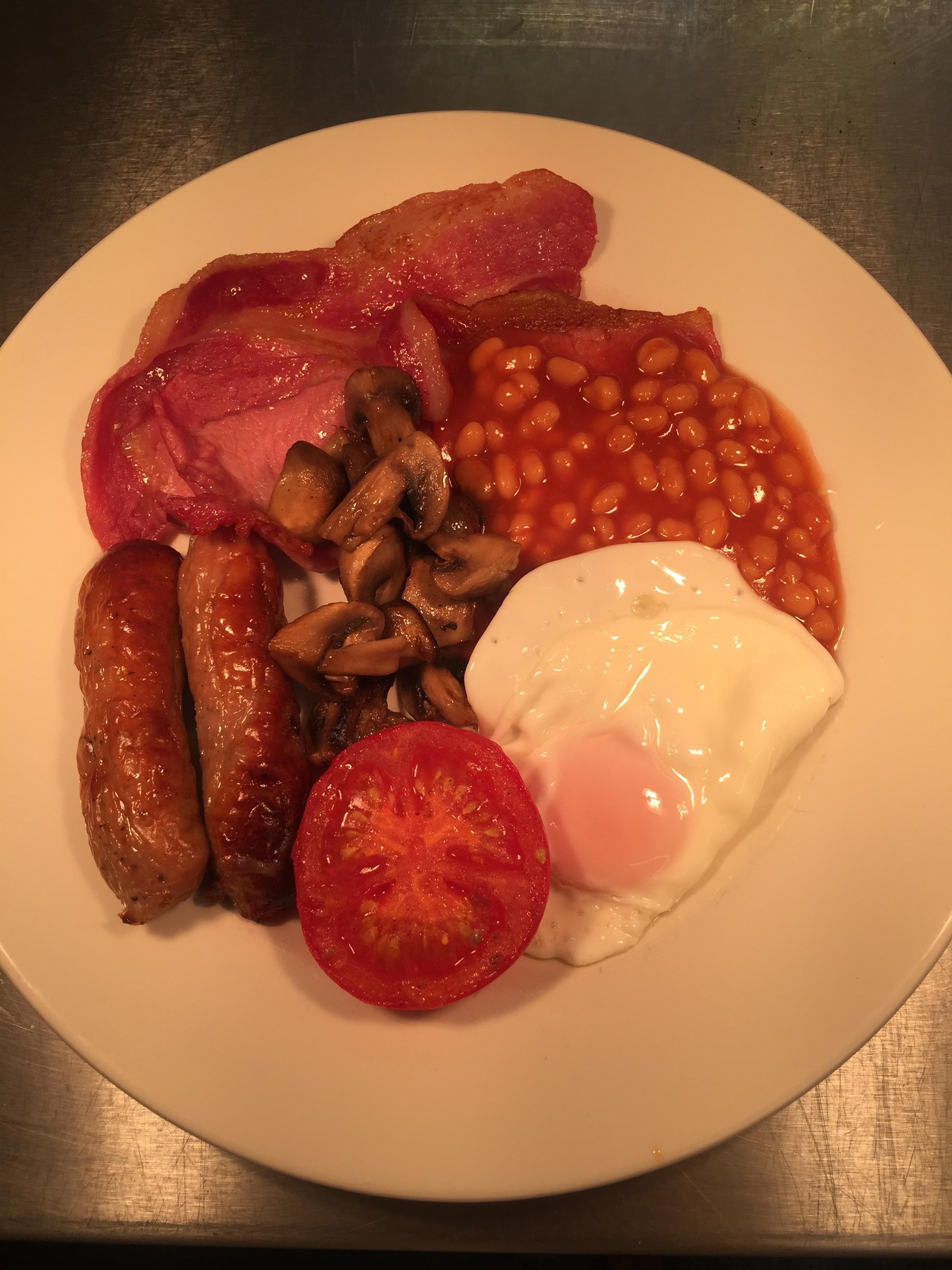 Un piatto colmo di cibo per la colazione: salsicce, pancetta, fagioli, funghi, un uovo fritto e una fetta di pomodoro.