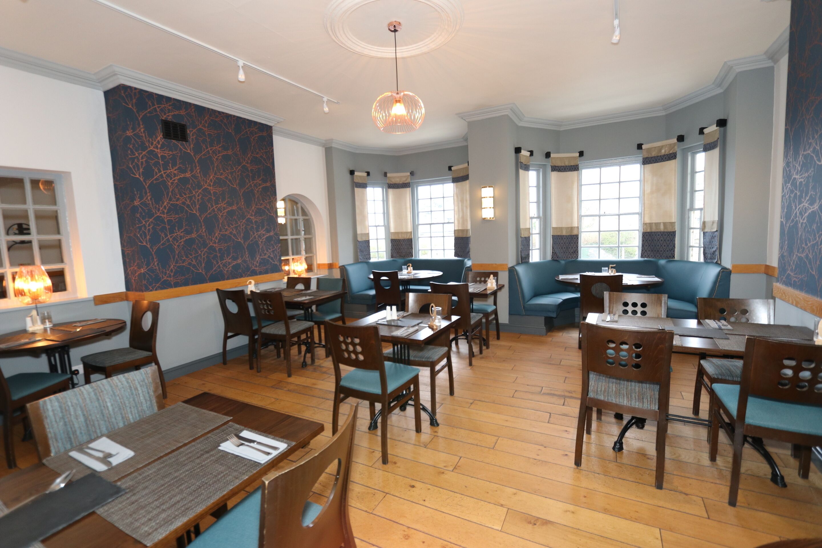 A restaurant with wooden tables and chairs, blue upholstered seating, and large windows with curtains.