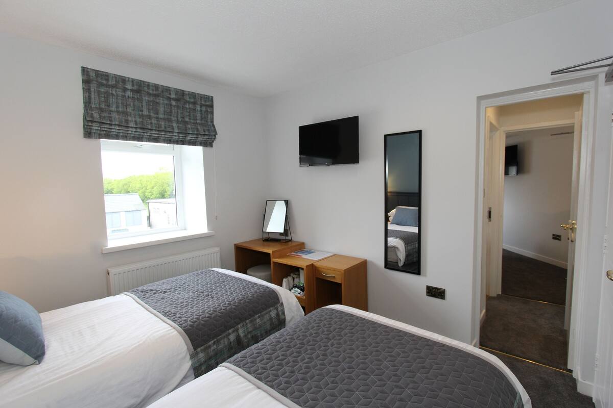 Ein Hotelzimmer mit zwei Betten, einem Schreibtisch mit Computer, einem Fernseher und einem Fenster mit Blick auf Gebäude.