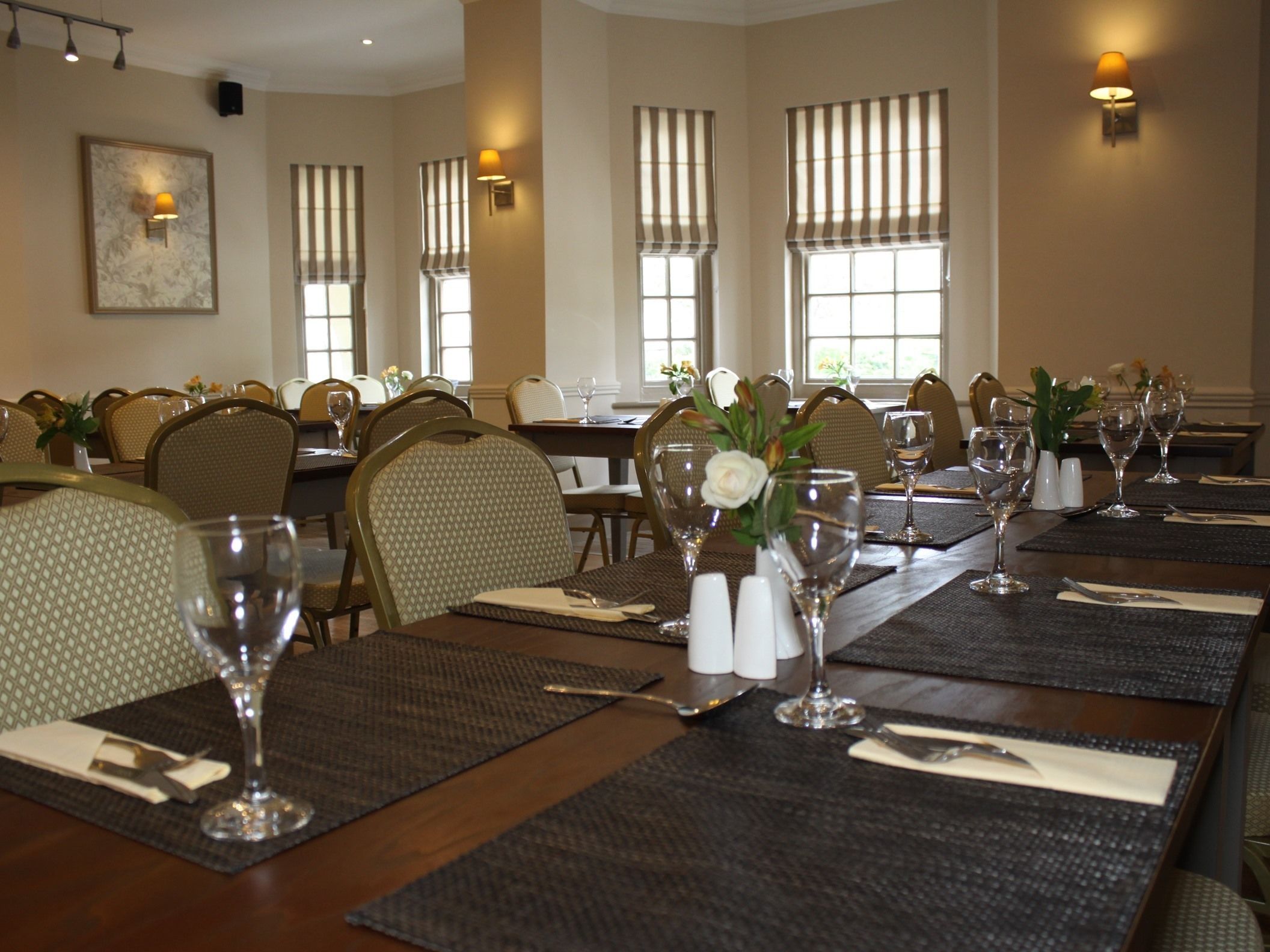 Une salle à manger avec plusieurs tables dressées pour le service, chacune agrémentée d’un vase de fleurs, de salières et de poivrières, ainsi que de verres transparents.