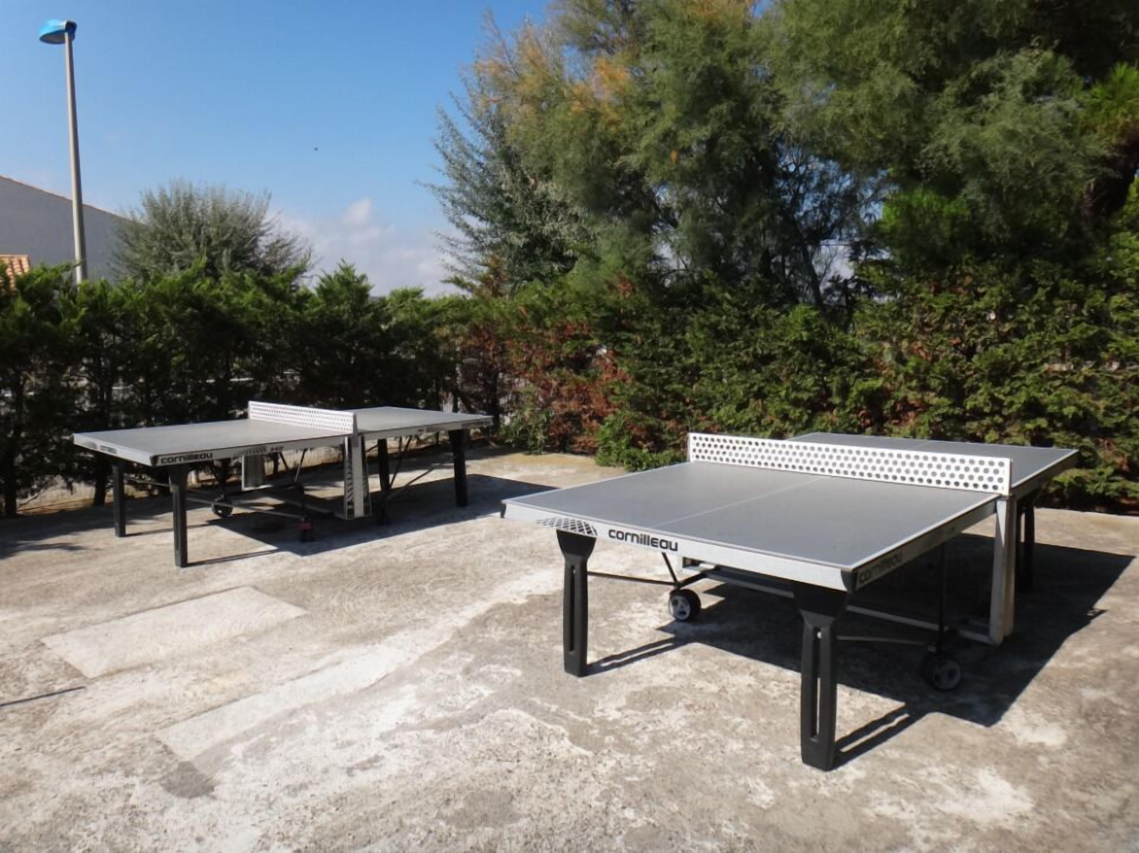 Deux tables de ping-pong sur roulettes, installées à l’extérieur sur une surface en béton, entourées de verdure.