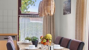 A dining area with a wooden table, cushioned chairs, a woven pendant light, a fruit basket, and a small potted plant.