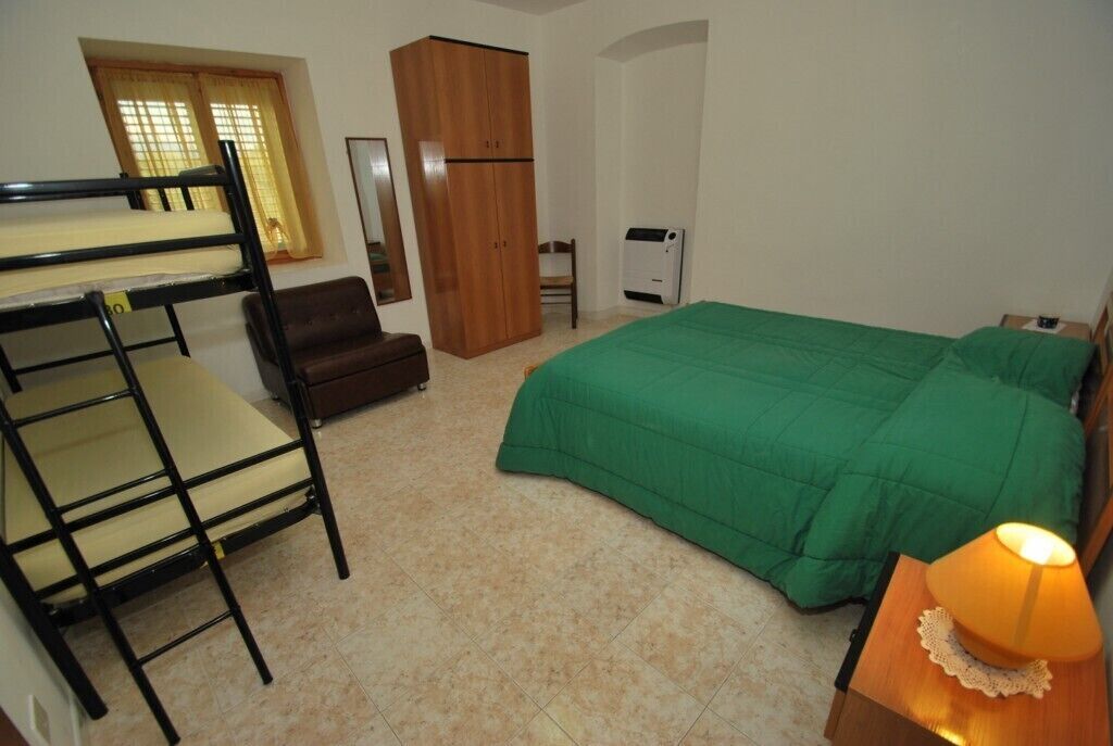 A bedroom with bunk beds, a green bedspread, a wooden wardrobe, a chair, a desk with a lamp, and a radiator.