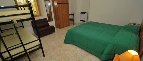 A bedroom with bunk beds, a green bedspread, a wooden wardrobe, a chair, a desk with a lamp, and a radiator.