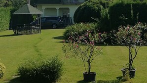 A large house with a green roof, a balcony, and a car parked in the driveway. There are trees and a lawn in the foreground. - Pine Wood Apartment 7 (Zalakaros)