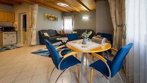 A dining area with a round table, blue chairs, a sofa, and a kitchen in the background. - Pine Wood Apartment 7 (Zalakaros)