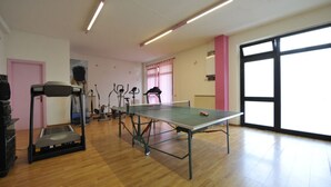 A room with a ping pong table, exercise equipment, and large windows.