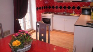 Een kleine keuken met een fornuis, oven en witte kastjes. Een rode tafel met een mand vol bloemen staat op de voorgrond.