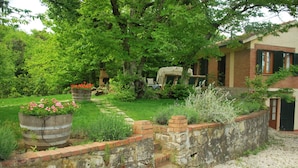 A rustic garden with a stone wall, a wooden barrel planter with flowers, and a brick pathway leading to a house with a chimney. - Villa La Quercia-Dalia (Val Canina)