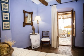 A room with a bed, a chair, a lamp, a mirror, and a door leading to an outdoor area with a patio and a view of a building.