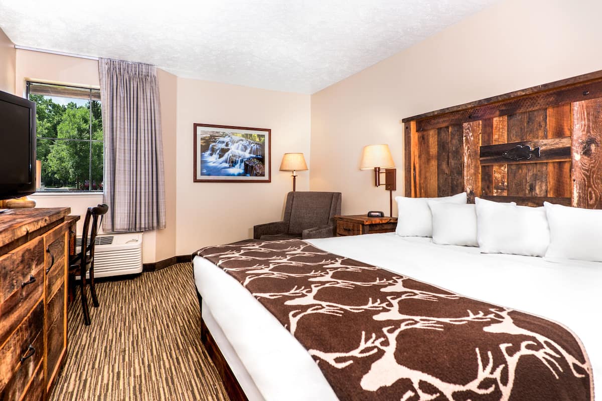A hotel room with a large bed, a wooden headboard, a television, a chair, a desk, and a window with a view of trees.