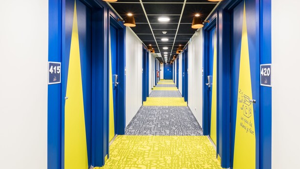 Ein Hotelgang mit blauen und gelben Wänden, nummerierten Türen und einem gemusterten Teppich.