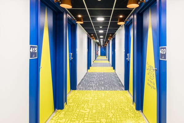 Un couloir d’hôtel aux murs bleus et jaunes, avec des portes numérotées et un tapis à motifs.