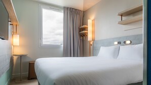 Un lit bien fait dans une chambre d’hôtel, avec une table de chevet, une lampe et une fenêtre avec des rideaux.