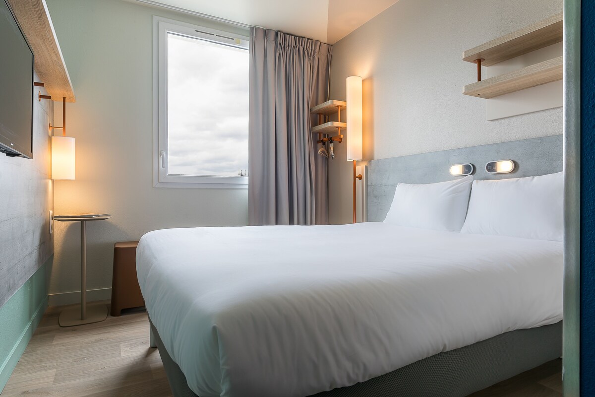 Un lit bien fait dans une chambre d’hôtel, avec une table de chevet, une lampe et une fenêtre avec des rideaux.