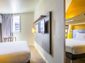 Une chambre d’hôtel avec deux lits, un téléviseur fixé au mur, une petite table et une fenêtre avec des rideaux.