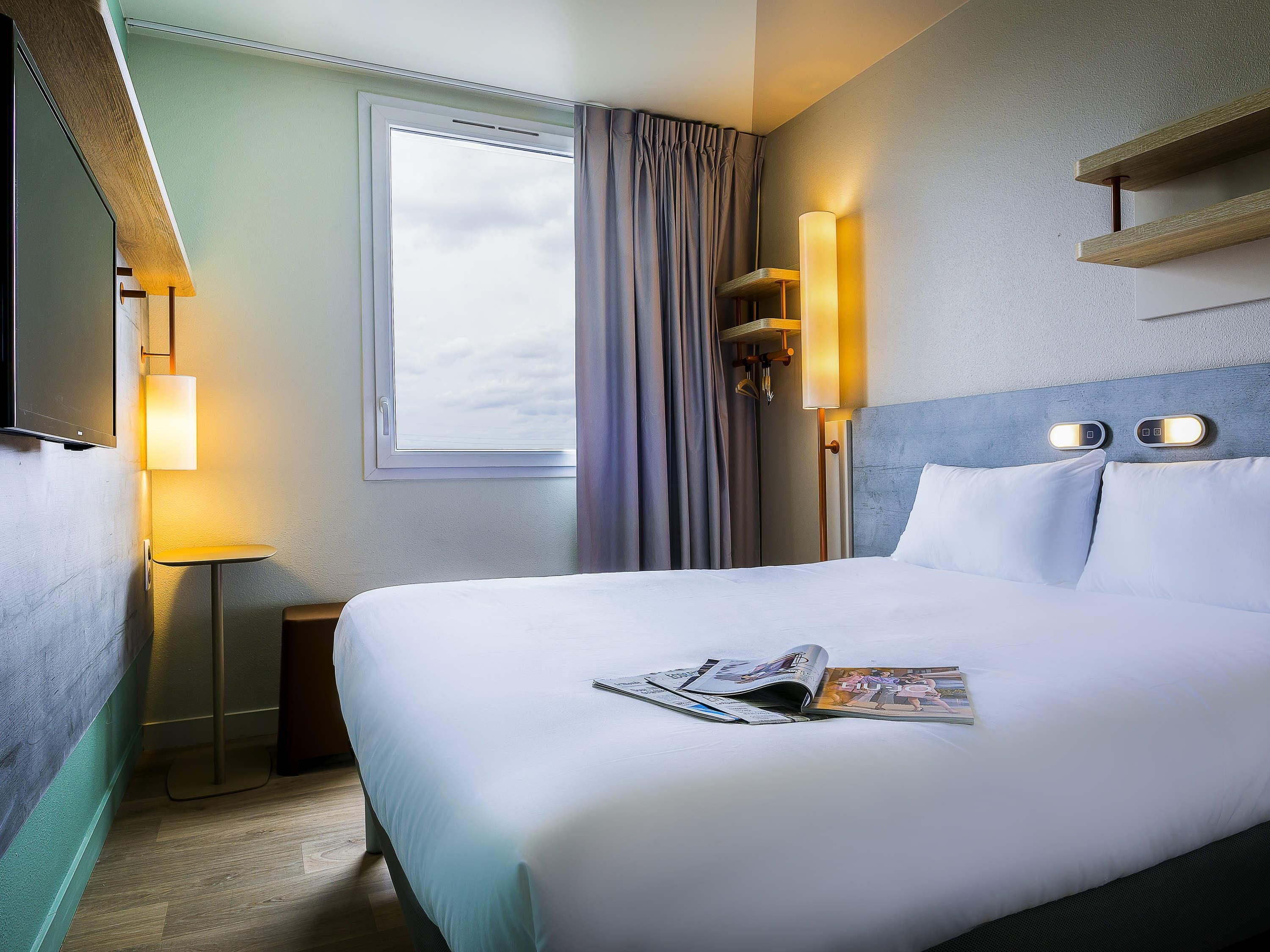 A hotel room with a bed, a bedside lamp, a window with curtains, a wall-mounted TV, and a magazine on the bed.