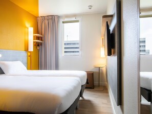 A hotel room with two beds, a window with a view of buildings, a flat-screen TV mounted on the wall, and a small table with a lamp. - ibis budget Paris Gennevilliers (Gennevilliers)