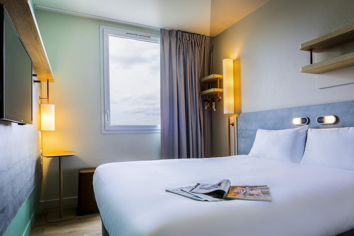 Een hotelkamer met een bed, nachtkastje, lamp, raam met gordijnen en een tv aan de muur.