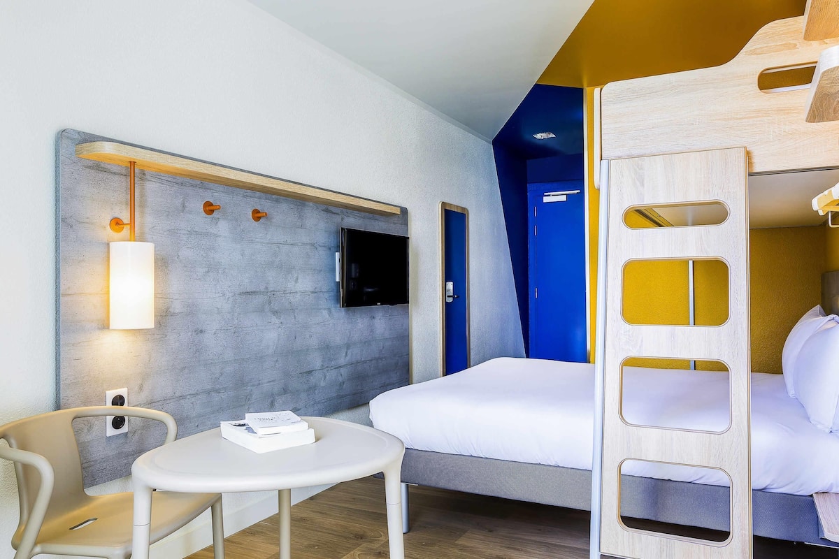 Een hotelkamer met een stapelbed, een kleine tafel met een stoel, een aan de muur bevestigde televisie, en een deur die naar een andere kamer leidt.