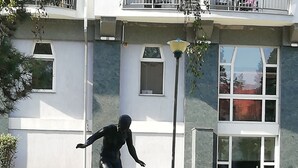 A statue of a person in a dynamic pose on a pedestal in a landscaped area with a building in the background.