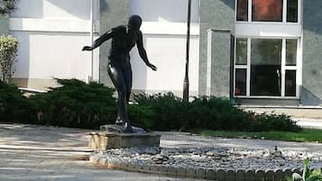 En staty av en person i en dynamisk pose, placerad på en piedestal i ett parkliknande område med en byggnad i bakgrunden.
