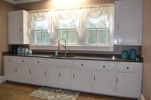A kitchen with white cabinets, a sink, and a window with curtains. - Starfish House Savannah Fort Stewart GA (Richmond Hill)