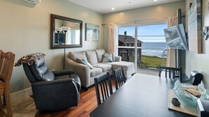 A modern living room with a sofa, a recliner, and a dining table. There is a large window offering a view of the ocean. - Three Arch Inn (Tillamook)