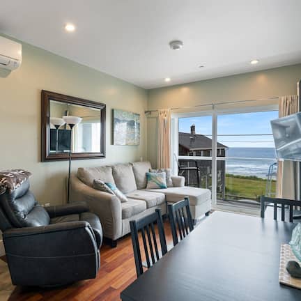 A modern living room with a sofa, a recliner, and a dining table. There is a large window offering a view of the ocean.