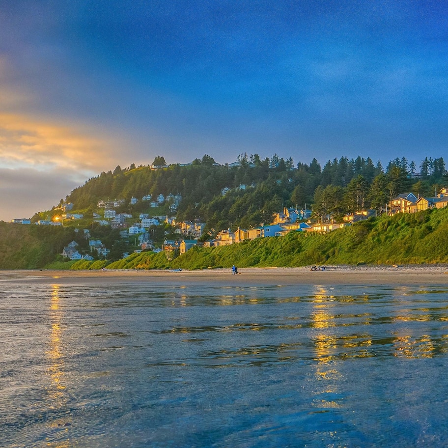 A coastal landscape with a beach, houses on a hillside, and a sunset.