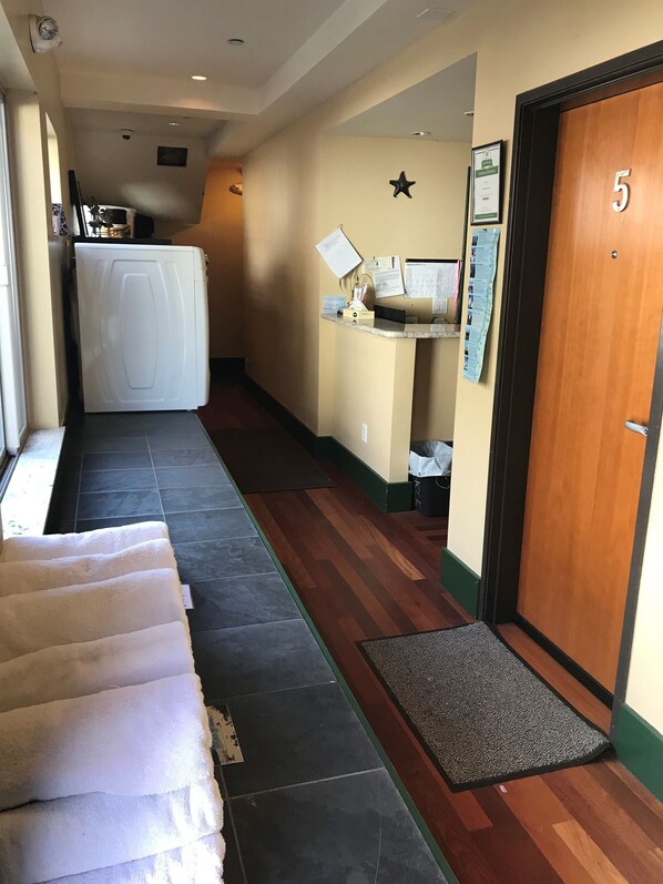 A hotel hallway with a reception desk, a wooden door marked with the number 5, a white washing machine, and a row of white towels. - Three Arch Inn (Tillamook)