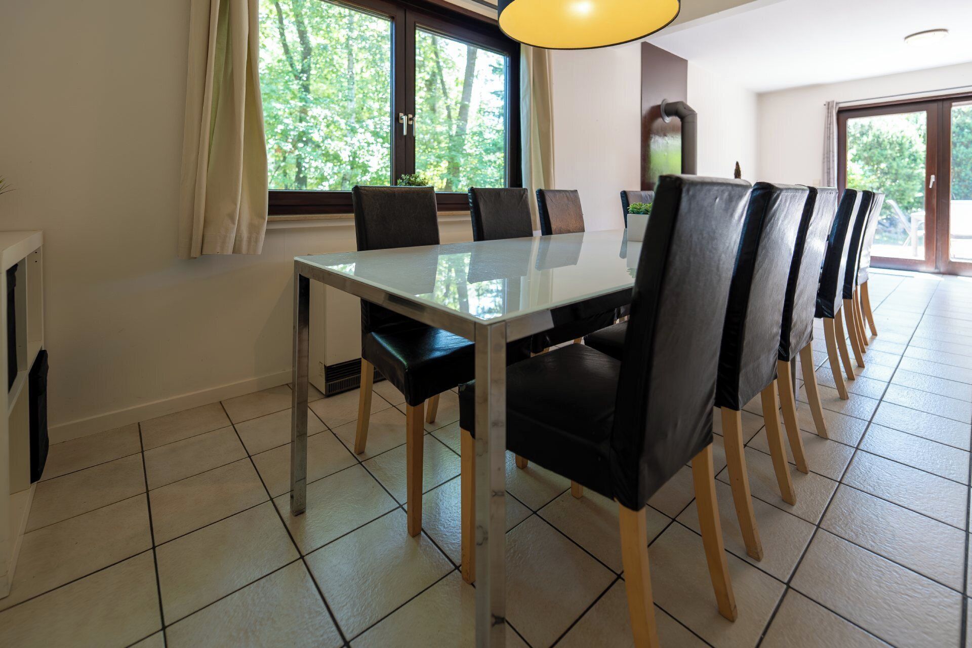 Een eetgedeelte met een glazen tafel en zwarte stoelen, grote ramen met gordijnen, en een betegelde vloer.