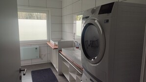 Een kleine wasruimte met een voorlader wasmachine, een klein wastafeltje en een raam.