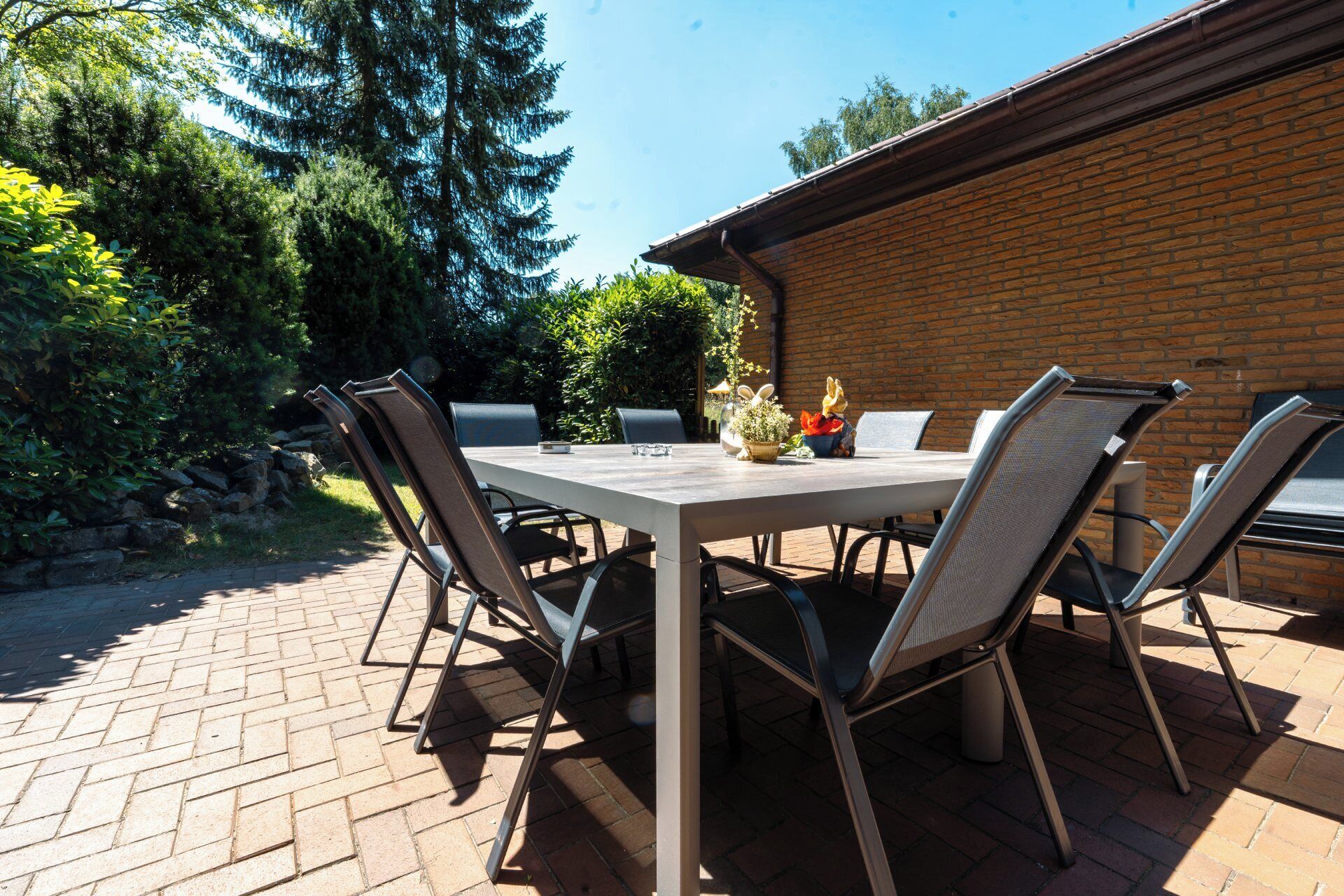 An outdoor patio with a rectangular table and four chairs, surrounded by a brick wall and greenery.