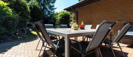 An outdoor patio with a rectangular table and four chairs, surrounded by a brick wall and greenery.