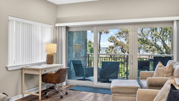 A modern room with a desk, office chair, and a sofa. There is a large sliding glass door leading to an outdoor area with a view of trees and a blue wall. - 463 Captains Walk is a gorgeous villa with breathtaking views of the ocean. (Hilton Head)