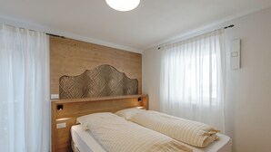 A room with a wooden headboard, two beds with striped bedding, white curtains, and a light switch on the wall. - Piz Boe' with private bio sauna (Livinallongo del col di lana)
