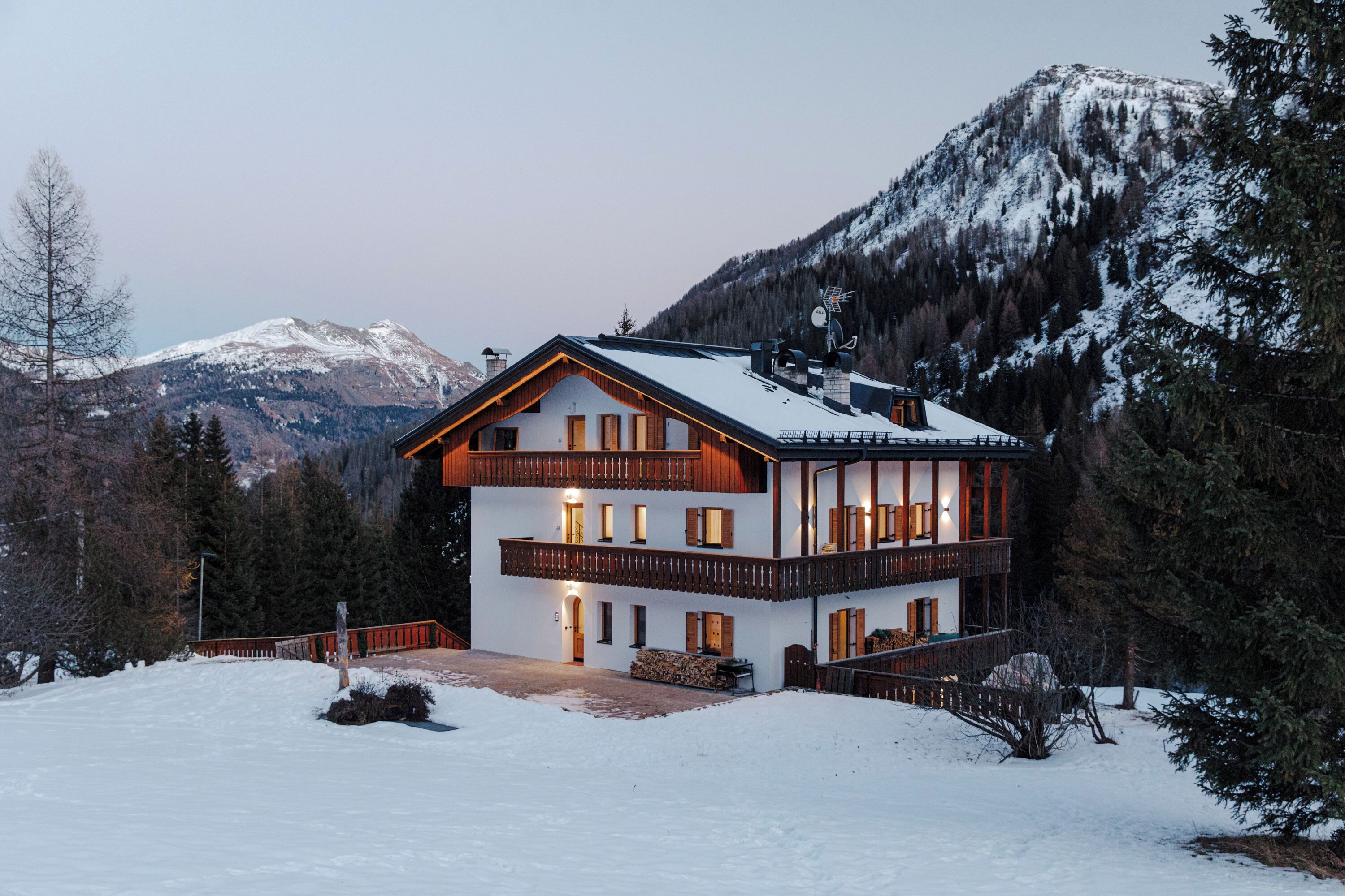 A two-story chalet with a balcony, surrounded by snow-covered mountains and trees.