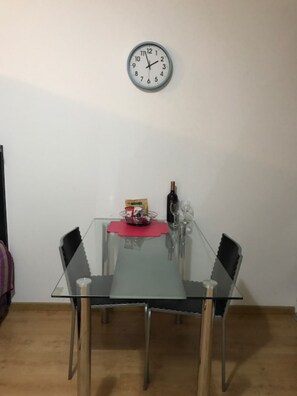Uma mesa de jantar com tampo de vidro, cercada por quatro cadeiras, e um relógio na parede acima.