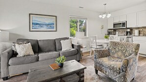 Un salón moderno con un sofá gris, un sillón estampado y una mesa de centro con una planta. Se ve un comedor con una mesa y sillas blancas, junto con una zona de cocina con armarios blancos y un microondas.
