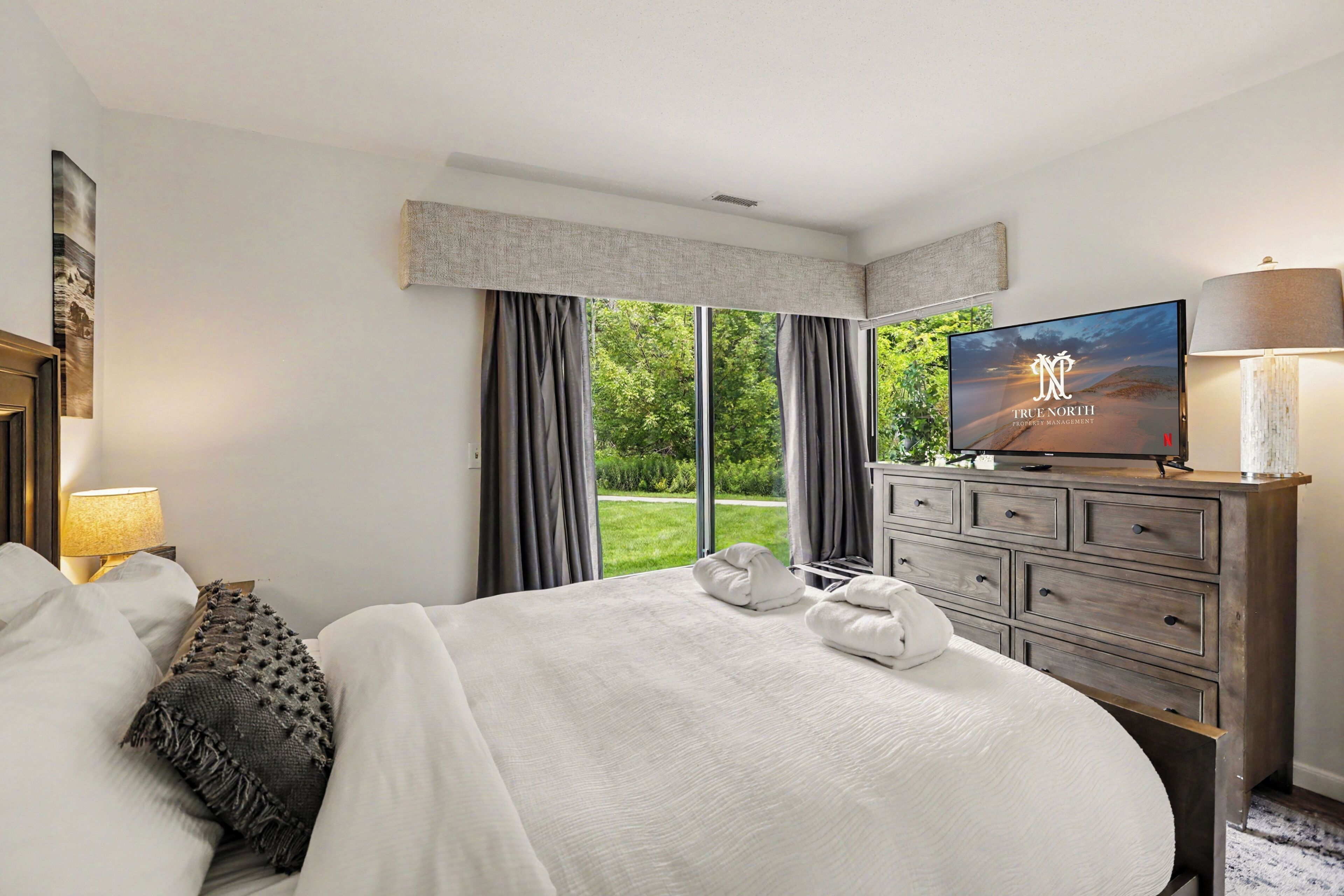 A hotel room with a large bed, a television, a dresser, and a view of greenery outside.