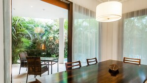 Un espace repas comprenant une grande table et des chaises, un luminaire suspendu et une vue sur un jardin luxuriant, visible à travers de grandes fenêtres.