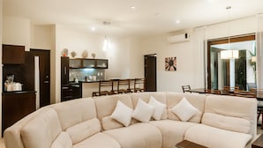 Un salon moderne avec un canapé d’angle beige, un coin repas avec des chaises en bois et une cuisine en arrière-plan.