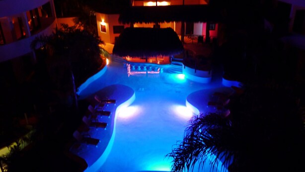 Une vue nocturne d’un complexe hôtelier avec une piscine illuminée de lumières bleues, entourée de palmiers et d’un bâtiment à toit de chaume.
